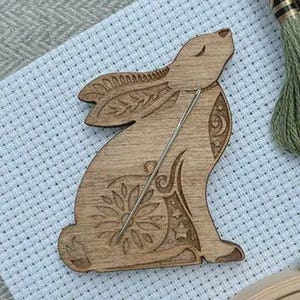 May include: A light brown wooden needle minder shaped like a sitting hare with laser-engraved floral, leaf, and star patterns. A thin silver metal needle is positioned diagonally across the center of the hare-shaped wooden surface.