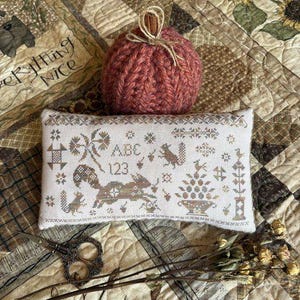 May include: A rectangular cream-colored fabric pincushion featuring intricate cross-stitch embroidery of squirrels, the letters ABC, the numbers 123, and floral motifs, paired with a round, ribbed, dark orange knit pumpkin topped with a cinnamon stick.