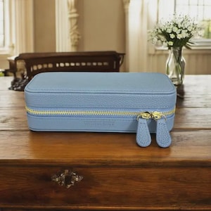 May include: A rectangular light blue jewelry case with a textured pebble grain finish. The case features a gold-toned metal zipper closure with two matching leather-like pull tabs. The brand name Winterbury is embossed on the front lower center.