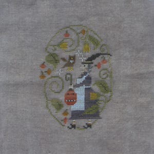 May include: A cross-stitch embroidery on gray fabric featuring a witch in a dark dress and white apron holding a red lantern. The design is framed by green vines, leaves, orange berries, and a small owl, all rendered in various colored threads.