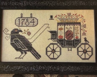 May include: A cross-stitch embroidery framed in black wood, depicting a black crow with a carriage pulled by two horses. The carriage is filled with pumpkins and has a sign that reads "1784".