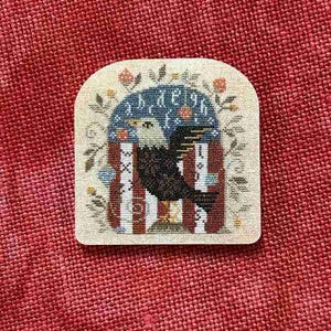 May include: A cross-stitch embroidery piece on a cream-colored fabric base featuring a black bird with a white head centered within an arch. The design includes a partial alphabet, vertical red and white stripes, and small floral accents in green and orange.