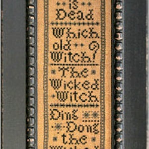 May include: A framed cross-stitch sampler with black embroidery on a beige background. The text reads "Ding Dong the Witch is Dead. Which old Witch? The Wicked Witch is Dead."