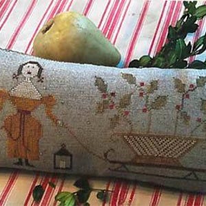 May include: A rectangular linen pillow featuring a cross-stitch design of a person in a yellow dress holding a lantern and pulling a sled filled with holly branches. The fabric is a light gray color with a decorative red stitched border.