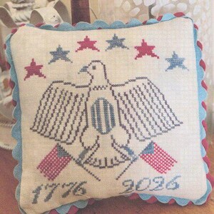 May include: A square cream-colored fabric pillow featuring a cross-stitched eagle with spread wings, flanked by two small flags. Above the eagle are five stars in red and blue. The text 1776 and 2026 is stitched below. The pillow has a scalloped blue and red trim.