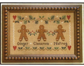 May include: Cross-stitch embroidery of three gingerbread people holding hands, with the text "Ginger Cinnamon Nutmeg" below. The design is framed in a brown wooden frame with a beaded border.