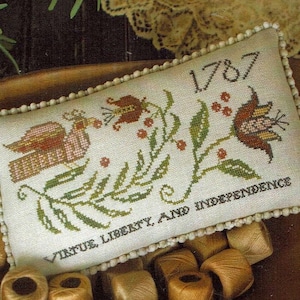 May include: A white pillow with a cross-stitch design featuring a bird, flowers, and leaves. The pillow has a white beaded trim and the text "1787 Virtue Liberty and Independence" is stitched on the pillow.