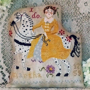 May include: A decorative, cross-stitched pillow featuring a woman on a spotted horse. The woman wears a yellow dress, and the pillow includes the text "I do," "Martha," and the numbers "12 59." The design incorporates floral and scrollwork elements.