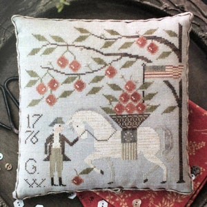 May include: A cross-stitched pillow with a light blue background features a horse carrying a basket of red cherries. A man in colonial attire stands beside the horse. The design includes an American flag and the year "1776".
