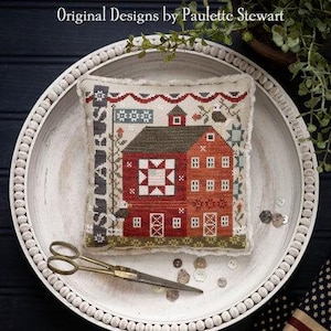 May include: A cross-stitch sampler featuring a red barn with a white star design, a flag, and the word "STARS". The sampler is displayed on a white tray with a beaded edge, along with scissors and buttons. The text "PLUM STREET SAMPLERS" is at the top.