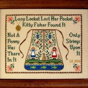 May include: A framed cross-stitch sampler on cream fabric featuring a central pocket illustration with two figures, a cat, and floral vines. The text reads: Lucy Locket Lost Her Pocket, Kitty Fisher Found It, Not A Penny Was There In It, Only Strings Upon It, LE 2011.