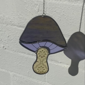 May include: A stained glass mushroom suncatcher with a purple cap and a yellow stem with a floral pattern. The suncatcher is hanging from a silver chain.