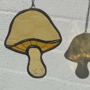 May include: A stained glass mushroom suncatcher with a yellow cap and brown stem hanging from a chain against a white brick wall.