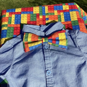 Può includere: Una camicia azzurra con bottoni e un papillon colorato. Il papillon e il tessuto sotto la camicia sono decorati con un motivo di colorati mattoncini giocattolo. La camicia è abbottonata e ha un colletto.