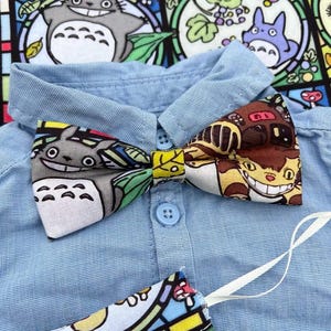 Peut inclure: Une chemise bleu clair à boutons avec un nœud papillon orné d'un imprimé de personnages de dessins animés colorés. Le nœud papillon présente des créatures grises et brunes souriantes. Un tissu assorti au motif vitrail est visible en dessous.