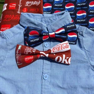 May include: Two bow ties, one with a Coca-Cola pattern and the other with a Pepsi pattern. The bow ties are on a light blue button-down shirt.