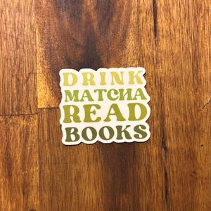 Könnte beinhalten: Ein Aufkleber mit dem Text "DRINK MATCHA READ BOOKS" in einer Retro-Schriftart. Die Wörter sind in Grün- und Gelbtönen gehalten.