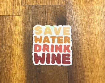 May include: A square sticker with the text "SAVE WATER DRINK WINE" in a gradient of orange and red colors. The sticker is on a wooden surface with a brown wood grain.