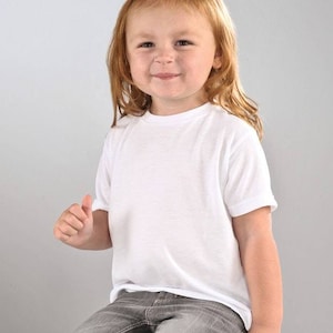 以下が含まれることがあります： 白い半袖Tシャツとグレーのジーンズを着た幼い子供。 子供は座って笑顔で、親指を立てています。 背景は無地のライトグレーです。