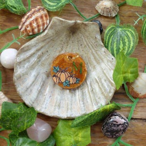 May include: A circular pendant with a glossy orange finish featuring a hand-painted illustration of three pumpkins and the letter M in green. The pendant has a small gold-toned metal loop at the top for attachment, showcasing a detailed autumn theme.