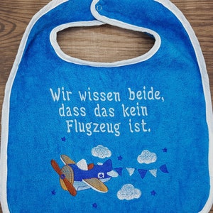 Könnte beinhalten: Blauer Baby-Lätzchen mit weißem Rand und gesticktem Text in Deutsch: "Wir wissen beide, dass das kein Flugzeug ist." Das Lätzchen zeigt ein weiß gesticktes Flugzeug mit Wolken und Sternen.