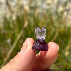 May include: A small, carved amethyst crystal figurine in the shape of a person wearing a dress. The crystal is clear and has a purple hue.