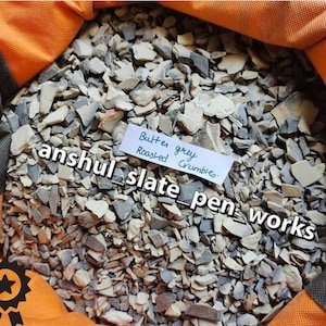 Op de afbeelding: Een close-up van een stapel kleine, gebroken stukjes leisteen. De leisteen is lichtgrijs van kleur en heeft een licht ruwe textuur. De tekst "Butter grey Roasted Crumbles" staat op een klein papiertje.