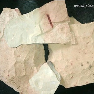 May include: A collection of irregularly shaped, light-colored slate pieces. The slate has a rough, textured surface with variations in color, including off-white and pale pink. The image includes the text "anshul_slate_pen_works".