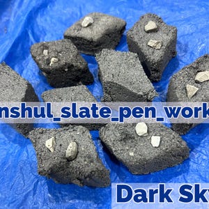 Pode incluir: Uma coleção de oito  costas de ardósia em forma de losango, de cor cinza escuro com manchas brancas. Os  costas estão dispostos sobre uma superfície azul. O texto "anshul_slate_pen_works" e "Dark Sky" é visível na imagem.