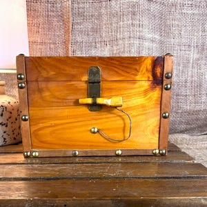 May include: A small, rectangular wooden box with a hinged lid and a metal clasp. The box is stained a warm brown color and has brass-colored studs along the edges. A small, decorative wooden handle is attached to the front.