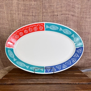 May include: Oval serving platter with a white center and a decorative border. The border features red, teal, and blue sections with geometric and fish-like patterns. The platter is placed on a wooden surface.