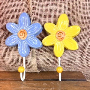 May include: Two ceramic flower-shaped wall hooks. One is light blue, the other is yellow, both with a white hook and a yellow ball at the end. The hooks are mounted on a wooden surface with a burlap background.