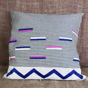 May include: Square decorative pillow with a gray and white striped pattern. The pillow features horizontal bands of blue, pink, and white rectangles. A white band with a blue zigzag design accents the bottom.