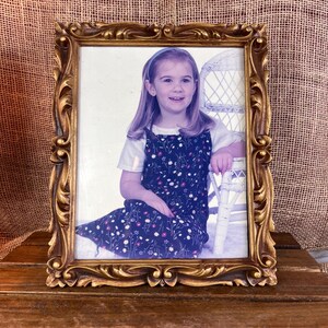 May include: Ornate gold-colored picture frame with a photograph of a young girl. The frame has detailed scrollwork and a rectangular shape. The girl is wearing a blue floral dress and a white shirt.