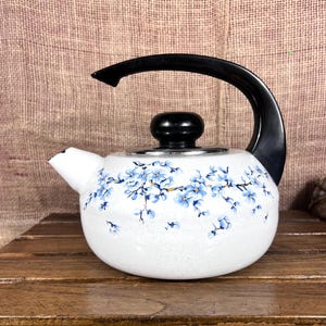 May include: White ceramic teapot with a black handle and lid. The teapot is decorated with a blue floral pattern. The spout is white, and the teapot sits on a wooden surface.