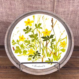 May include: A round decorative tray with a wooden frame. The tray's center features a floral design with yellow flowers and green leaves on a white background. The tray is supported by a metal stand and rests on a wooden surface. The text "Goodwood" is printed on the tray.