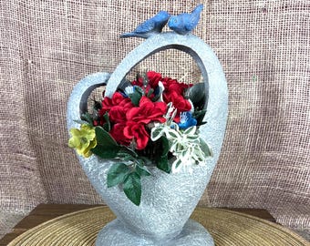 May include: A gray, heart-shaped decorative vase with a handle, filled with red, white, and blue flowers. Two blue bird figurines perch on the handle. The vase sits on a round, woven placemat.