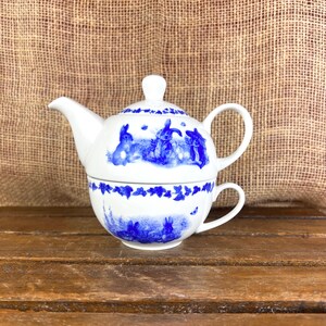 May include: A white ceramic tea-for-one set featuring a teapot stacked on a teacup. The set is decorated with blue illustrations of rabbits and ivy leaf borders. The teapot has a lid with a knob handle, a curved spout, and a side handle.