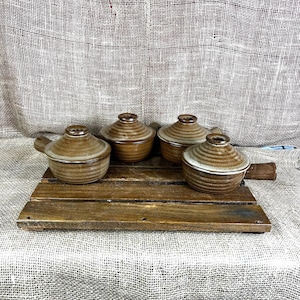 May include: Four small brown ceramic pots with matching lids and side handles, arranged on a rectangular wooden board. Each pot features a circular, ribbed design on the lid and a smooth, glazed finish in varying shades of earthy brown and tan.