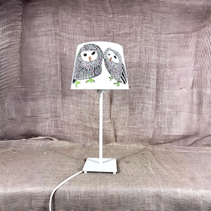 May include: A white table lamp with a lampshade featuring two owl illustrations. The owls are depicted in black and gray with green feet, set against a white background. The lamp has a square base and a slender stem.