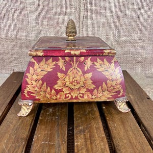 May include: A decorative box with a red background and gold floral design. The box has a gold finial on the lid and gold feet. The box is rectangular with curved sides.
