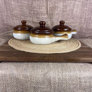 May include: Three ceramic soup bowls with lids. The bowls are white with a tan band and brown lids. One bowl has a handle. The bowls are on a wooden tray with a woven mat.