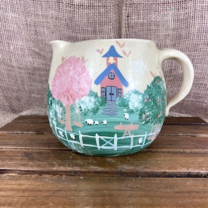 May include: Cream-colored ceramic pitcher with a hand-painted pastoral scene. The design features a pink tree, a blue-roofed building, and a white picket fence. The pitcher has a handle and a spout, and the artist's signature is visible.