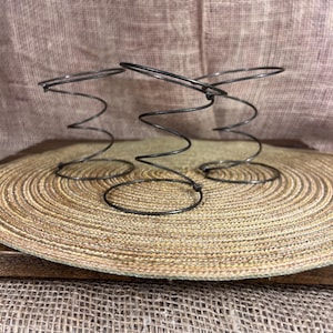 May include: A close-up of a metal spring, likely from a mattress or furniture, with a spiral design. The spring is dark gray and sits on a round, woven mat. The background is a textured, tan fabric.