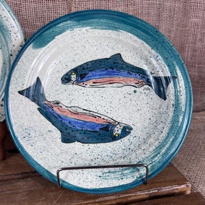 May include: A ceramic plate with a hand-painted design of two fish. The fish are blue with pink and blue stripes, set against a speckled white background. The plate has a teal blue rim and is displayed on a metal stand.