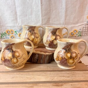 May include: Set of four ceramic mugs with a brown and beige glaze. Each mug features a hand-painted design of a bunch of grapes.