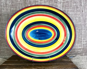 Puede incluir: Una bandeja ovalada de cerámica que presenta una serie de anillos concéntricos en colores vibrantes, incluyendo amarillo, rojo, verde y azul oscuro. El diseño irradia desde un centro azul sólido, creando un patrón multicolor en la superficie.