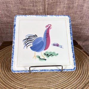 May include: Square ceramic plate with a rooster illustration. The rooster is blue and red, with a black tail and yellow legs. The plate has a white background with a blue border and a small barn illustration.