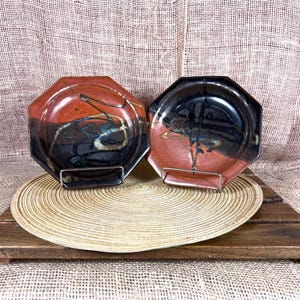 May include: Two octagonal ceramic plates with a unique glaze. The plates feature a blend of red, black, and blue hues with abstract designs. They are displayed on metal stands, resting on a woven placemat and a wooden surface.