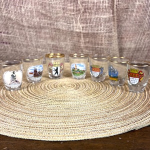 May include: A collection of seven clear shot glasses with gold rims, each featuring a different city crest or emblem. The glasses are arranged on a round, woven placemat. The shot glasses are a collectible item.